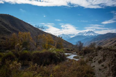 Küçük bir nehri ve köy yolu olan dağ vadisi. Uzakta mavi sisli, karla kaplı tepeleri olan bir dağ silsilesi. Parlak sonbahar gökyüzü Güzel bulutlar ve sarı ağaçlarla.