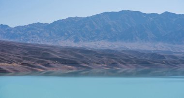 Uzakta mavi sisli dağları olan cansız bir vadinin ortasında turkuaz bir göl. Parlak gölü olan gri, donuk dağlar..