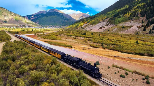 Tren turístico con carbón dejando pequeña ciudad a lo largo del río en las montañas Fotos de stock
