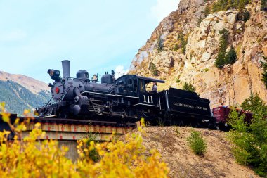 Lokomotif treni sonbahar renkleri ve kayalık dağlarla köprüden geçiyor