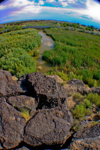 Large black rocks obscure the mouth of a river that flows through green pastures in a fish-eye shot — Stock Photo, Image