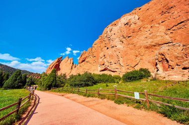 Mavi gökyüzüne karşı yeşil alanda yürüyen yolun yanındaki kırmızı kayalar duvarı.