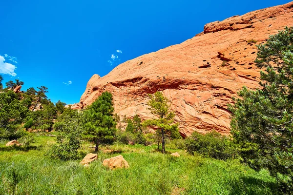Parede de penhasco de rocha vermelha ao lado de campo verde exuberante de grama e árvores — Fotografia de Stock