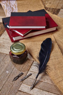 Ek ucu olan siyah tüy kalemi kapalı bir mürekkep tenceresi ve üç kırmızı ve siyah derginin yanında yatıyor.