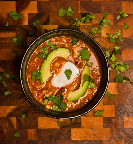 Birds eye view of an orange creamy chicken enchilada soup with avocado sour cream and cilantro on a cutting board