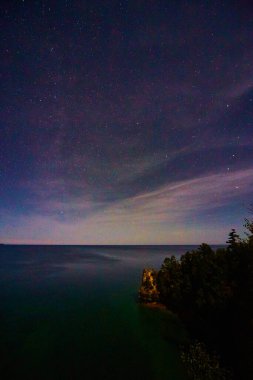 Michigan gölünün üzerinde yumuşak pembe bulutların fısıltılarıyla gece gökyüzü ve sağ altta karanlık bir orman olan bir uçurum.
