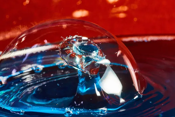 Bubble on blue water surface with two water drops colliding in air on red background — Stock Photo, Image