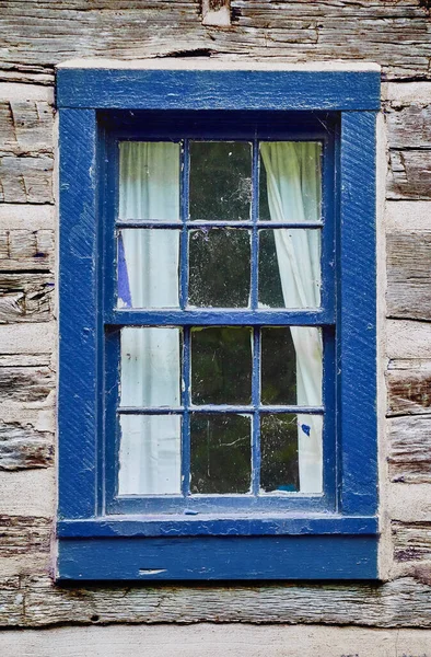 Old faded light wood building with a dark blue wooden window frame and white curtains pulled back