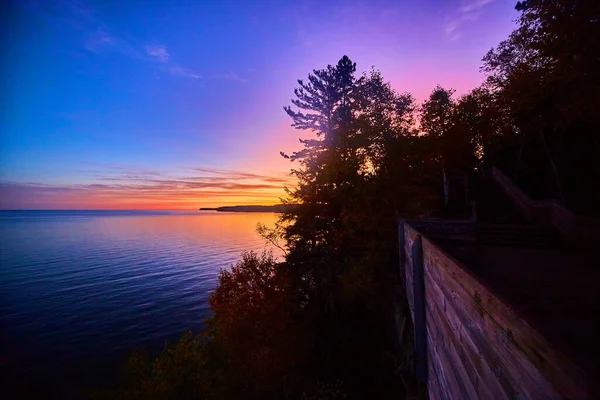 Восход солнца из золота, красного, фиолетового, розового и синего над озером или океаном с деревянной пешеходной дорожкой справа, которая проходит через лес — стоковое фото