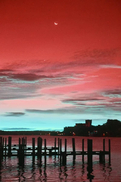 Infrarossi di banchine vuote sagoma in un fiume viola e verde acqua con un cielo rosso e viola con una luna crescente e cielo nuvole blu all'alba — Foto Stock