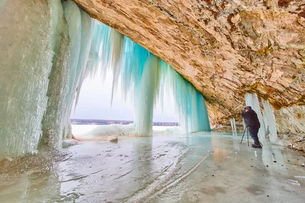 Fotograf w jaskini lodowej na zamarzniętym jeziorze rejestrując obraz dużego zamarzniętego wodospadu i niebieskich sopli — Zdjęcie stockowe