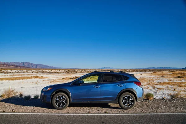 Blue Subaru Crosstrek 'in görüntüsü yolda park edilmiş ve arka planda Ölüm Vadisi' nin beyaz kum çölü var.