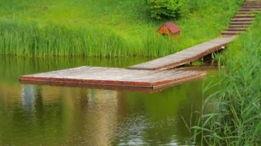 Tahta bir iskeleye zum yap, gölün üzerinde ahşap basamaklar, rıhtımın yanında ahşap bir kabin, kıyı boyunca sazlıklar ve çimenler büyür. Yaz, gün, 4K