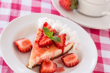 Taze dilimlenmiş çilekli cheesecake, kahve fincanı ve kırmızı kareli kumaş.