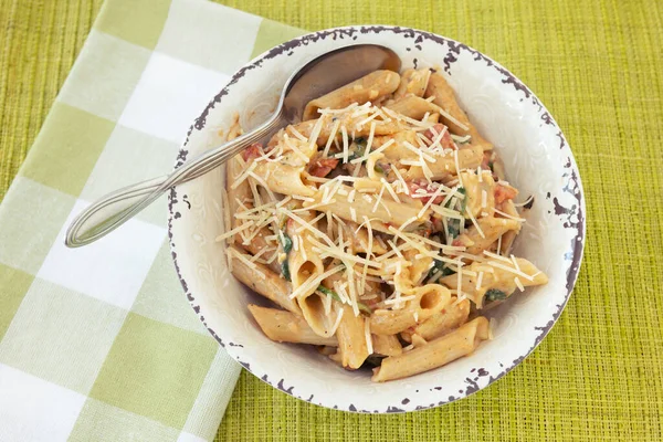 Gluten free penne pasta dinner with cheese, spinach, chicken, tomatos with spoon in rustic bowl on green placement with green napkin