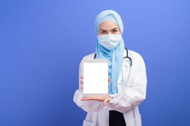 Türbanlı kadın bir müslüman doktor mavi arka plan stüdyosunun üzerinde tablet kullanarak cerrahi maske takıyor..