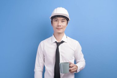 Beyaz kask takmış, mor arka plan stüdyosunun üzerinde kahve içen kendinden emin bir erkek mühendisin portresi.