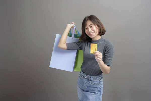 一张年轻美丽的亚洲女人的肖像 她拿着信用卡和彩色购物袋 隔着灰色的背景工作室 — 图库照片