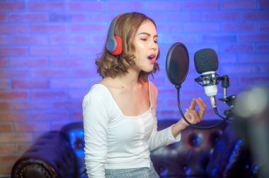 Renkli ışıklarla dolu bir müzik stüdyosunda şarkı söylerken mikrofonla kulaklık takan genç bir bayan şarkıcı..