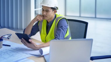 Ofiste tablet üzerinde çalışan beyaz kasklı bir erkek mühendis.