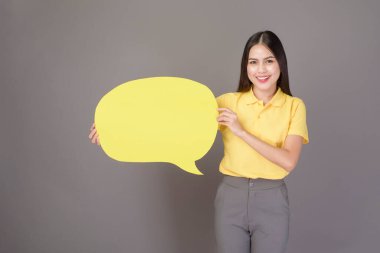 Sarı tişört giyen kendine güveni tam genç bir kadın gri arka plan stüdyosunda sarı boş bir konuşma yapıyor.