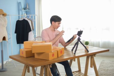 Asyalı bir adam dükkanında canlı yayında akıllı telefonun önünde giysilerini gösteriyor. Teknoloji çevrimiçi iş konsepti.