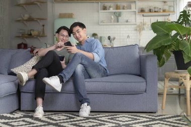 Genç, gülümseyen eşcinsel çift akıllı telefon kullanıyor ve evdeki oturma odasında dinleniyor, LGBTQ ve çeşitlilik konsepti.