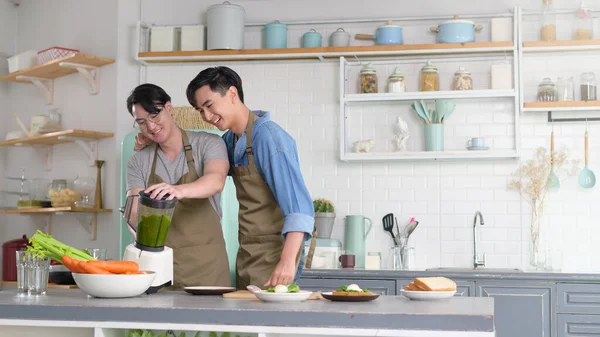 Genç, gülümseyen eşcinsel çift evde, mutfakta meyve suyu makinesiyle meyve suyu yapıyor, LGBTQ ve çeşitlilik konsepti.