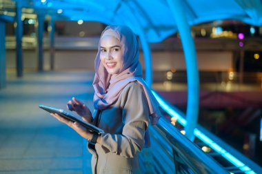Genç Müslüman bir iş kadını modern şehirde gece tablet bilgisayar kullanıyor, iş teknolojisi yaşam tarzı konsepti.