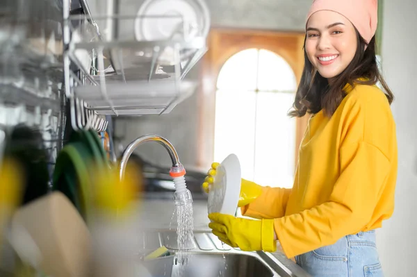 一位年轻快乐的女人 戴着黄色手套 在厨房里洗碗 — 图库照片