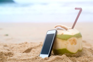 Kumsalda akıllı telefonlu pipet içen taze hindistan cevizi, seyahat ve tatil konsepti.