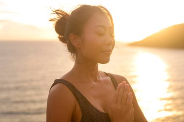 Spor giyim sektöründe genç bir Asyalı kadın gün batımı, sağlık ve meditasyon konsepti sırasında deniz kenarındaki kayada yoga yapıyor. 