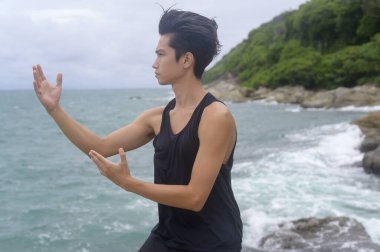 Spor giyimli genç adam dövüş sanatları yapıyor, Qigong, Tai Chi deniz kenarındaki kayada, sağlık ve meditasyon konsepti. 