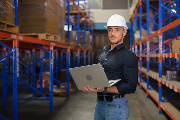 A Young caucasian male worker wearing helmet using laptop in modern warehouse.