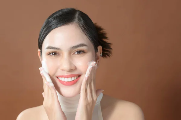 A young woman with beautiful face smiling, washing her face over brown background , beauty skin care concept