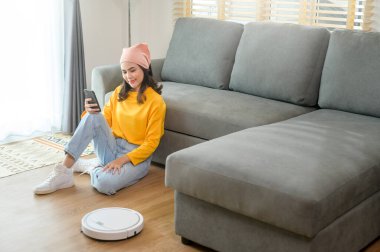 Genç ve mutlu bir kadın oturma odasında rahatlarken ve akıllı telefon kullanırken robot elektrikli süpürge çalışıyor.