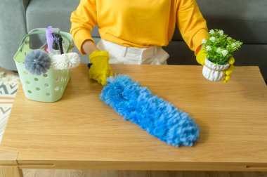 A Young happy woman wearing yellow gloves  and dusting the table in living room.