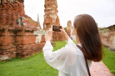 young beautiful woman wearing protective mask traveling and taking photo at thai historical Park, Holidays and cultural tourism concept.