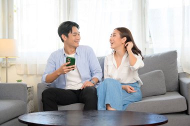 Genç, gülümseyen Asyalı çift evdeki oturma odasında akıllı telefon kullanıyor. 