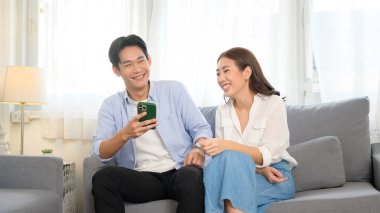 Genç, gülümseyen Asyalı çift evdeki oturma odasında akıllı telefon kullanıyor. 