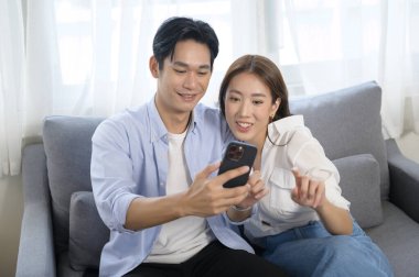 Genç, gülümseyen Asyalı çift evdeki oturma odasında akıllı telefon kullanıyor. 