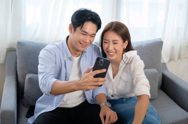 Genç, gülümseyen Asyalı çift evdeki oturma odasında akıllı telefon kullanıyor. 