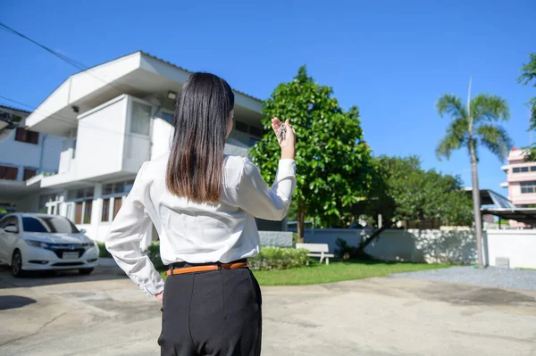 A Female real estate agent standing in front of the house and holding keys, Mortgage, loan, property and insurance concept.