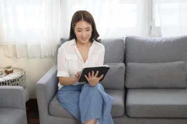 Genç Asyalı kadın oturma odasındaki kanepede tablet kullanıyor.