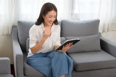 Genç Asyalı kadın oturma odasındaki kanepede tablet kullanıyor.