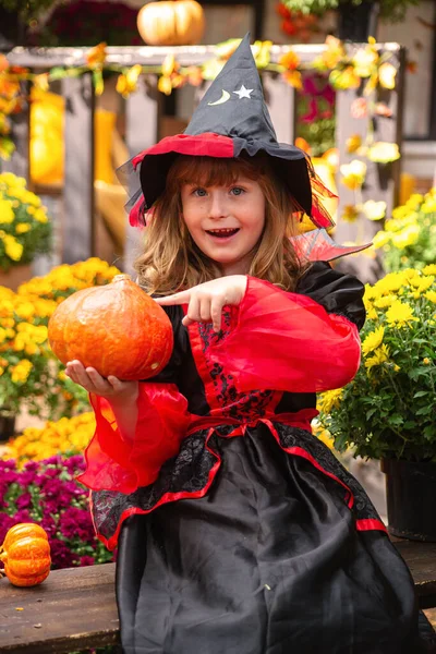 little girl dressed as a witch with a pumpkin celebrates Halloween in the fall in the garden near the house