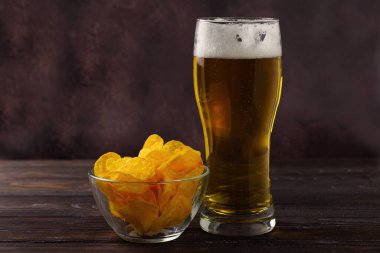 Bira ve patates cipsi. Büyük köpüklü bir bardak light bira. Doğal ahşap bir masada cam bir kasede kırıntılar. Yiyecek ve içecek.