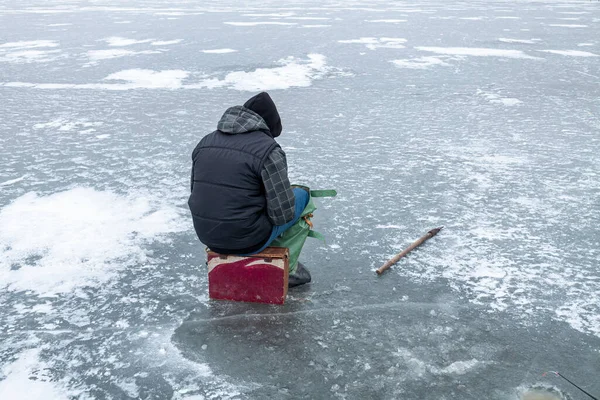 Buzda balık tutan bir balıkçı, balıkçı sandığına oturur ve donmuş bir gölde balık yakalar.