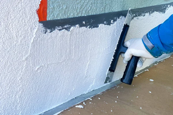 İnşaatçı evin ön cephesine yeni bir doku uyguluyor. İnşaatçının elindeki büyük spatula bir evin duvarlarını onarıyor.