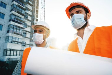 Çalışma kıyafetleri ve tıbbi maskeli iki adam nesne üzerinde planlar üzerinde çalışıyor.